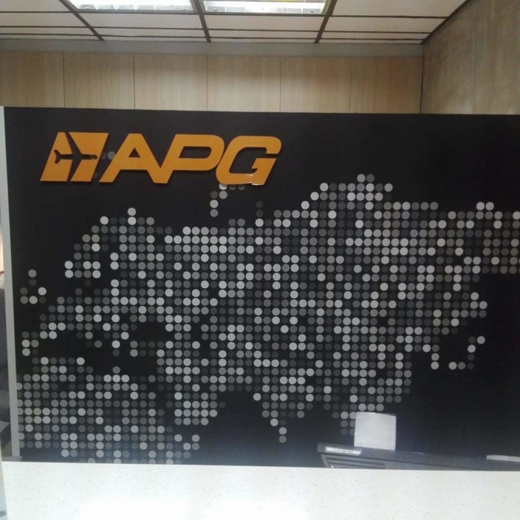 Acrylic signage with yellow APG logo and silver dots behind a white desk in Makati office, by M&G Global Ads.