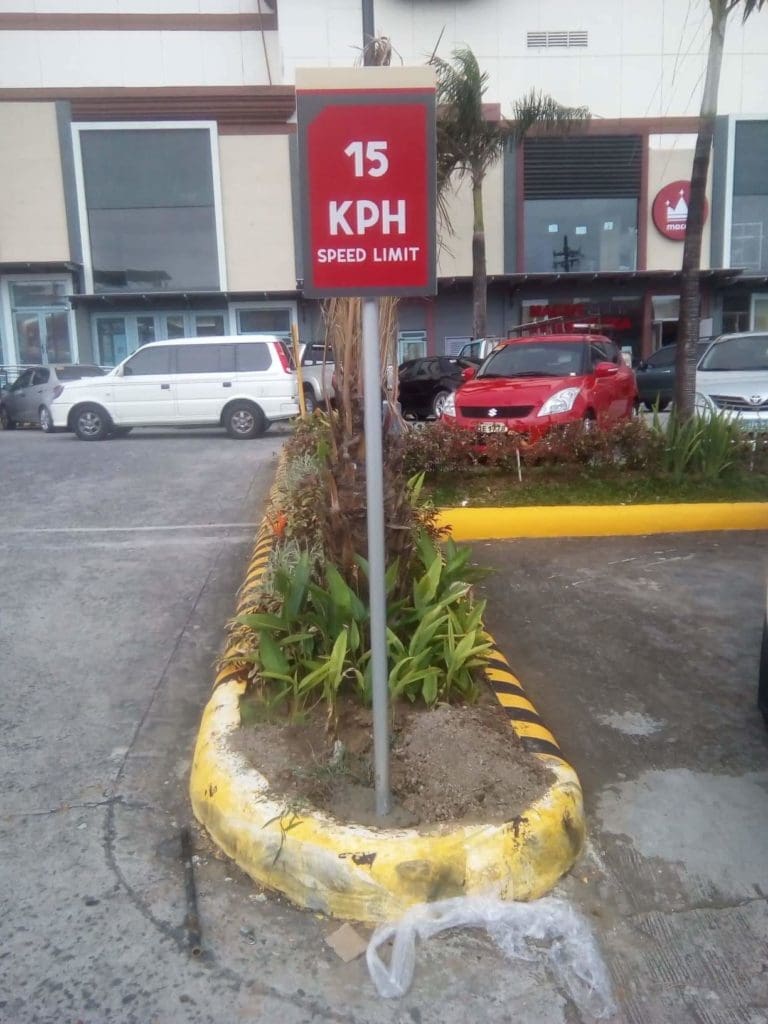 Parking lot speed limit sign 15 KPH in Makati, red and white metal signage by M&G Global Ads, cars and greenery in background.