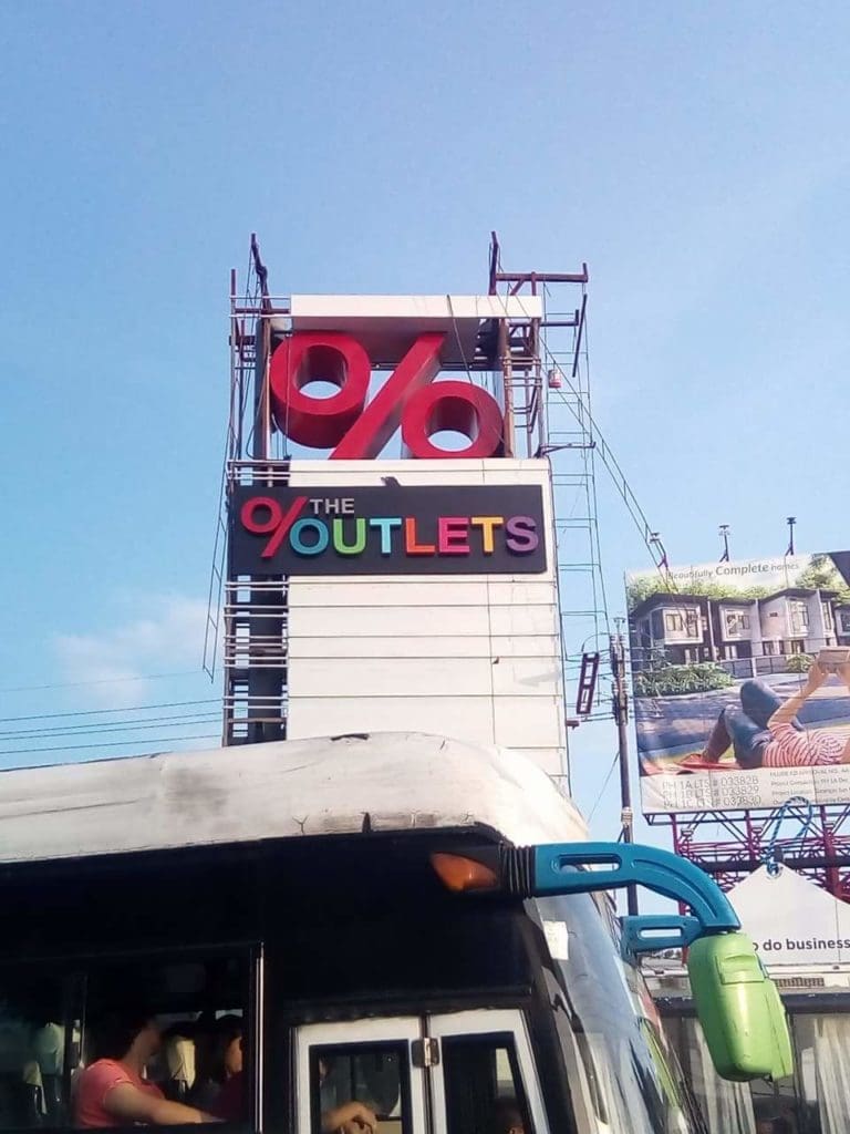 Outdoor mall signage for THE OUTLETS with red percent symbol in Ortigas, bus and billboard visible. By M&G Global Ads.