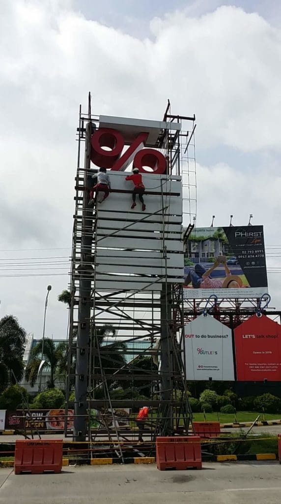 Workers installing a roadside billboard sign with red percent symbol in Quezon City, orange barriers visible, by M&G Global Ads.