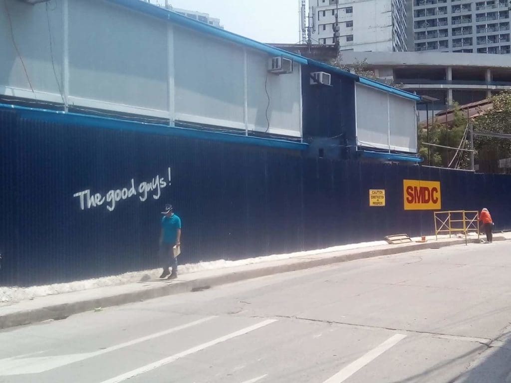 Two people sweep by blue construction fence with SMDC acrylic signage and The good guys! text in Makati by M&G Global Ads.