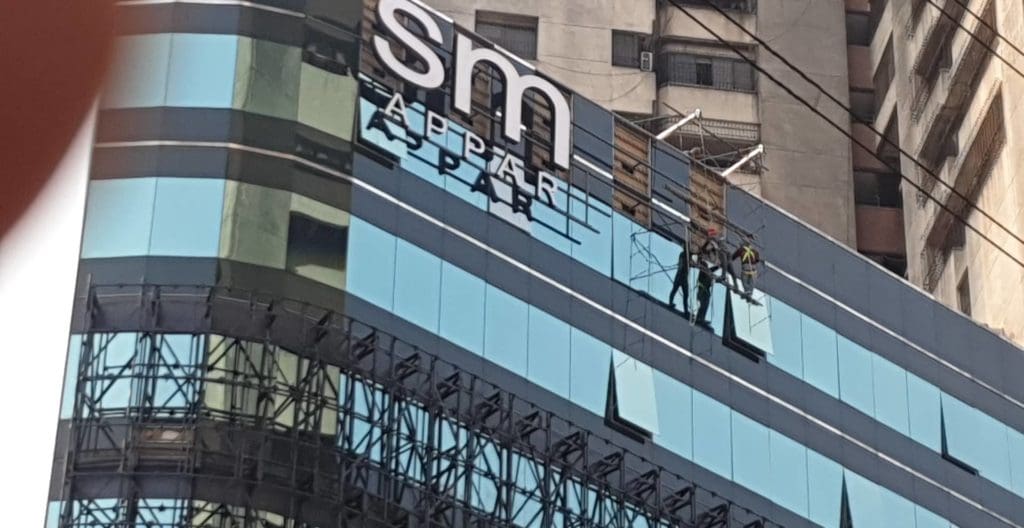 Acrylic signage installation for SM APART in Makati, two workers on glass building facade with ropes and cables by M&G Global Ads.
