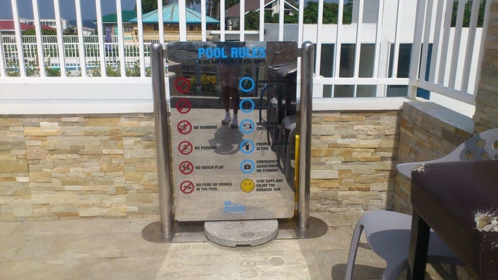 Pool rules metallic signage on a stone wall near pool in Mandaluyong, with chair, table, and sunlight reflections by M&G Global Ads.