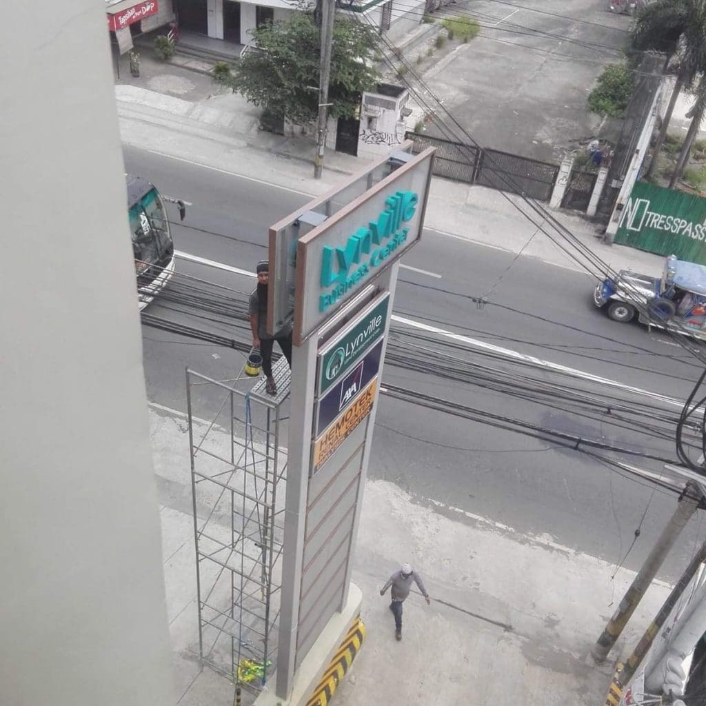 High-rise pylon signage installation for Lynville Residences in Quezon City, workers on scaffold by busy road, by M&G Global Ads.