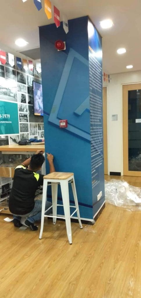 Installing acrylic display signage pillar in Makati; person kneels by base, with stool nearby. Indoor setup by M&G Global Ads.