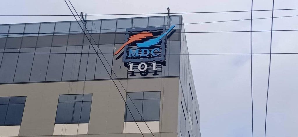 Metallic outdoor business signage for MDC with orange and blue dolphin logo in Makati, by M&G Global Ads. Power lines in view.