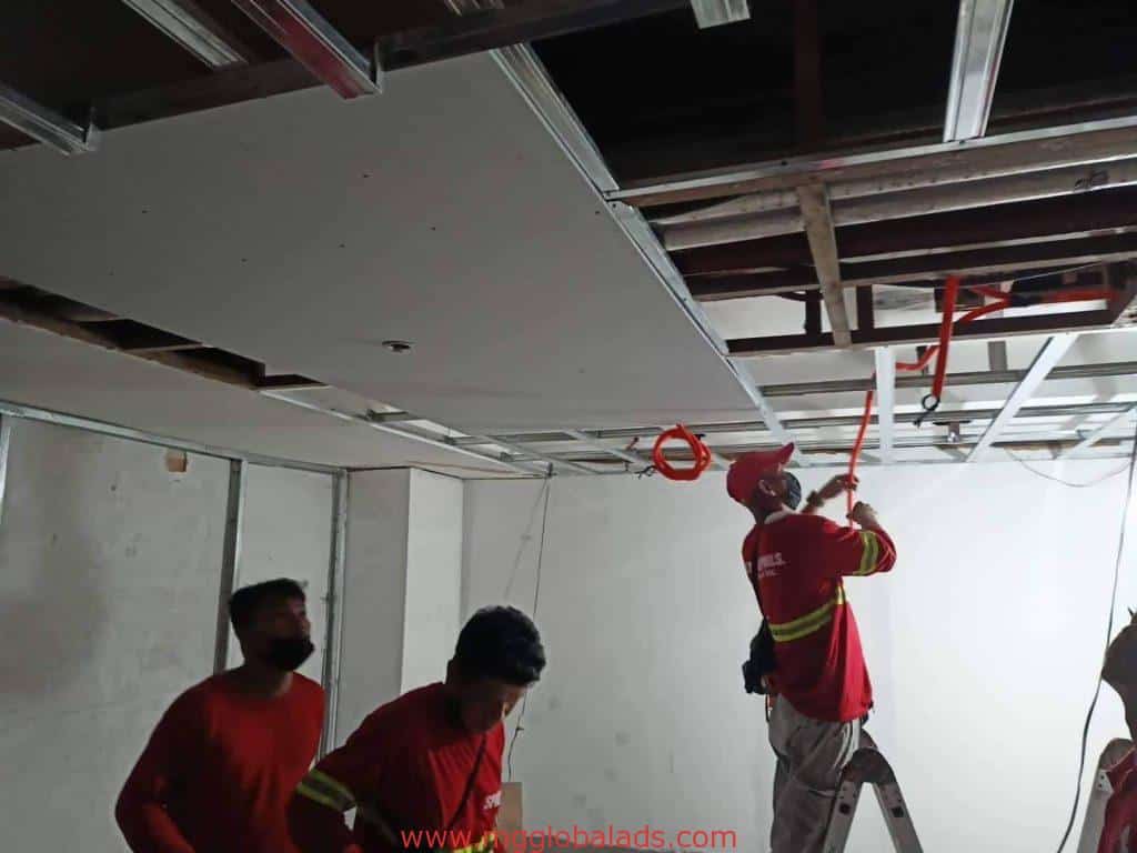 Workers installing drywall panels on ceiling with visible metallic sign in Makati office under construction by M&G Global Ads.