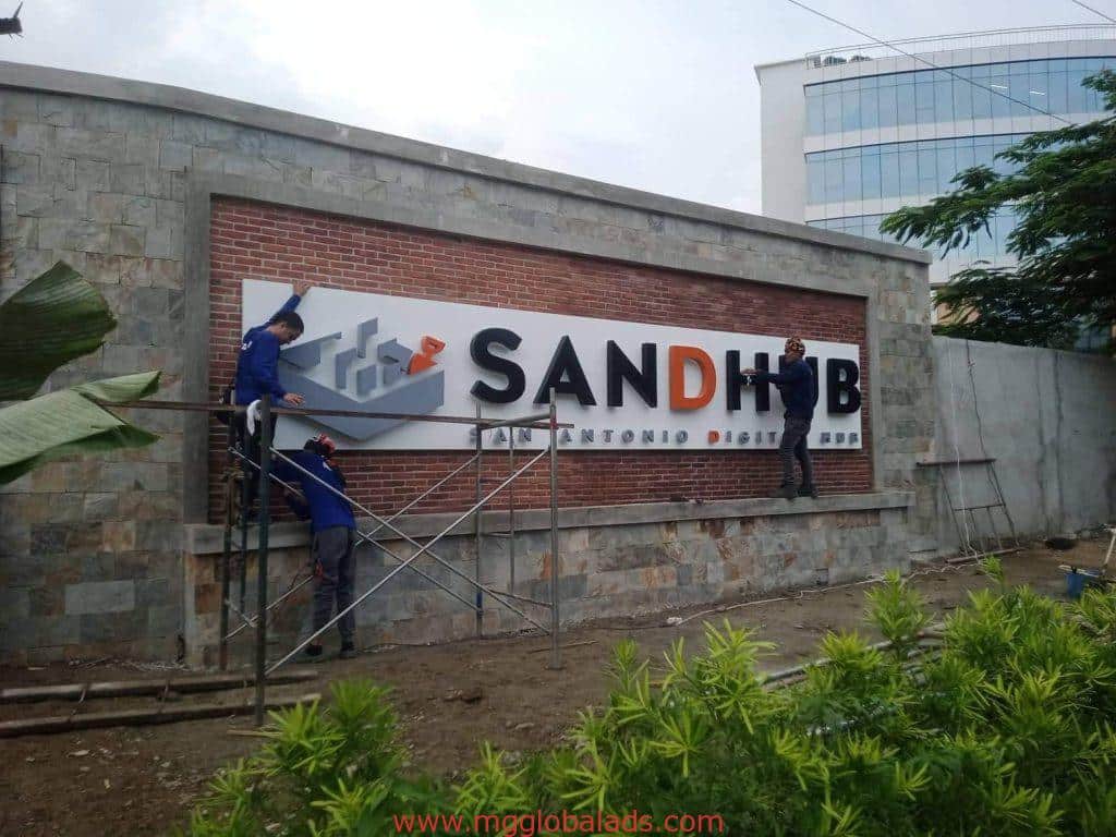 SANDHUB outdoor building signage installation by three workers in BGC on brick wall with scaffolding, by M&G Global Ads.