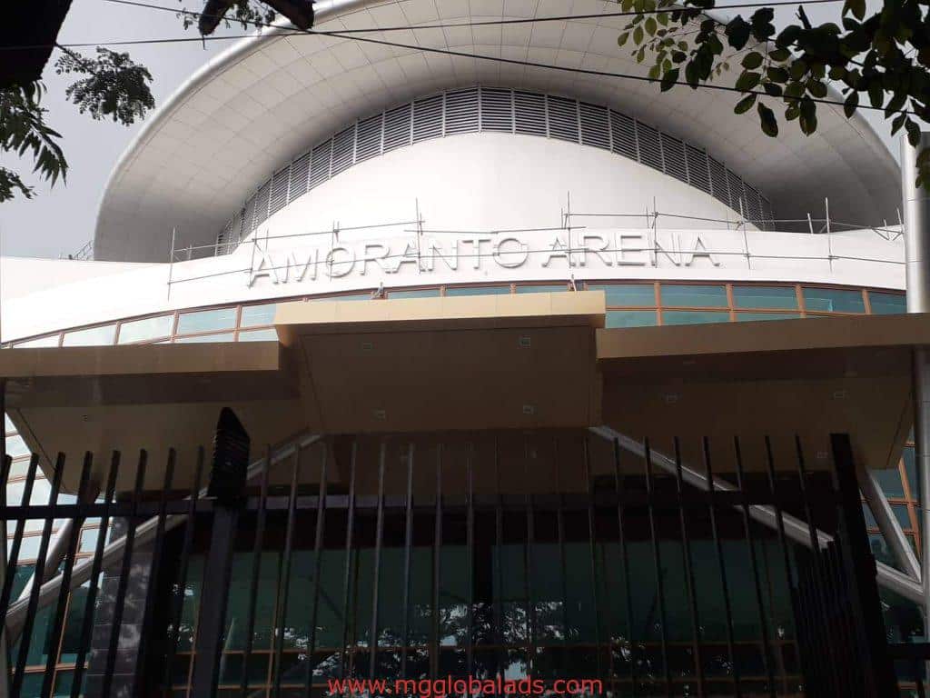 Amoranto Arena channel letter signage on a modern building in Quezon City, behind fence and trees, by M&G Global Ads.