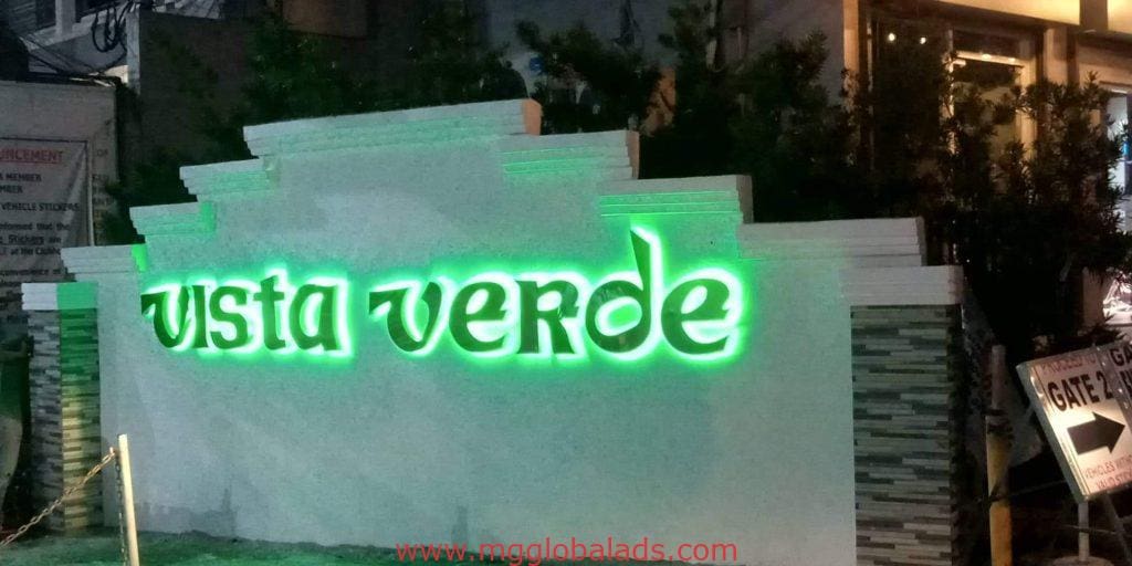 Outdoor neon signage for Vista Verde in bright green, surrounded by plants in Makati. Building background. By M&G Global Ads.