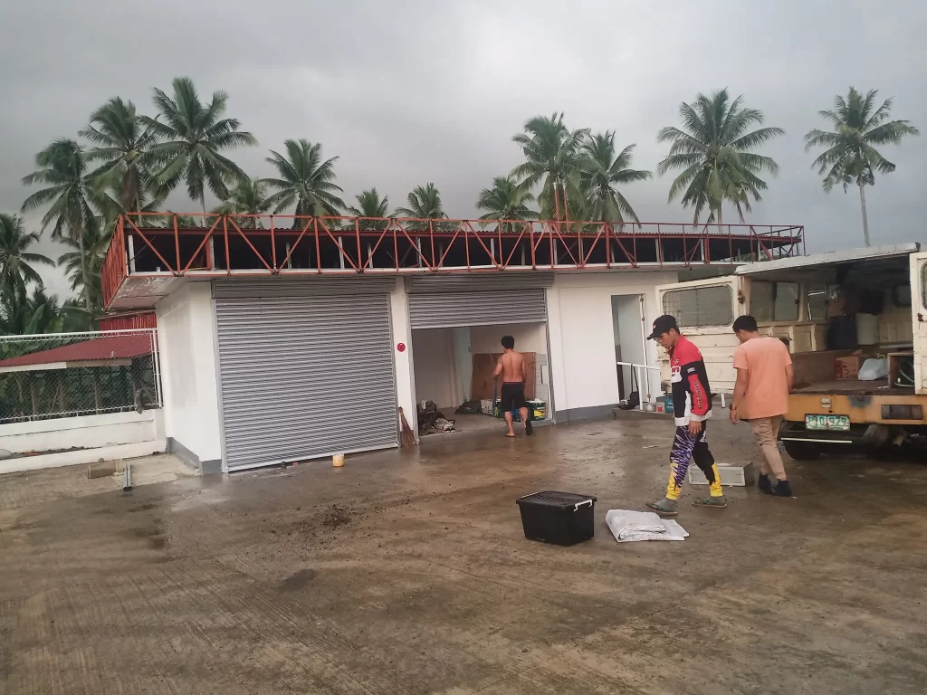 People clean outside a small building with metal shutters and retail signage in Quezon City; palm trees, supplies, truck by M&G Global Ads.