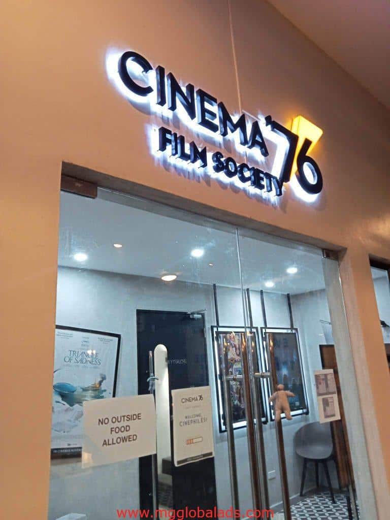 Cinema 76 LED signage at glass entrance in Quezon City with interior posters, chairs, and no outside food sign by M&G Global Ads.
