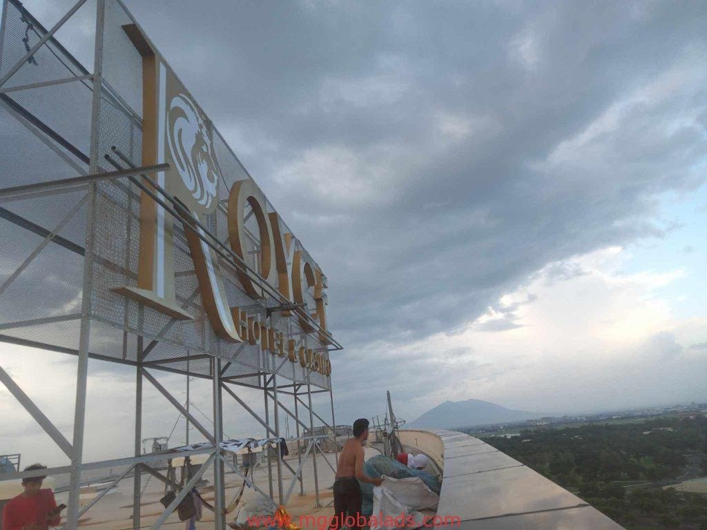 Workers install metallic building signage on a rooftop in Makati, mountain views in background, by M&G Global Ads.