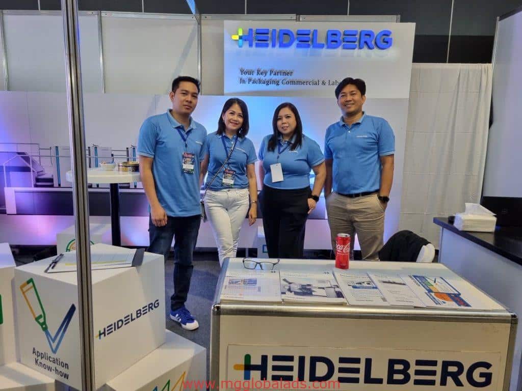 Four staff in blue shirts at a Heidelberg expo booth with metallic company signage in Makati by M&G Global Ads.
