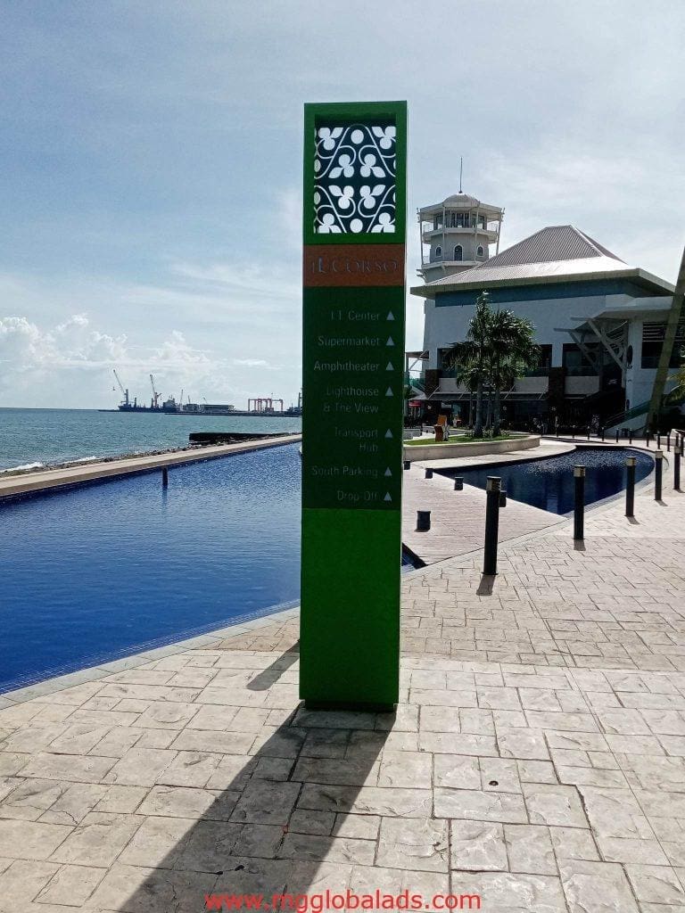 Acrylic wayfinding signage in Makati lists supermarket, amphitheater, lighthouse; near water feature. By M&G Global Ads.