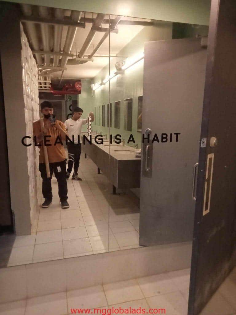 Acrylic signage CLEANING IS A HABIT in a bathroom mirror, two men reflected, selfie and mop. Makati. by M&G Global Ads.
