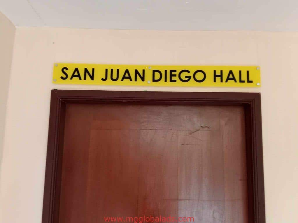 Yellow outdoor building signage for San Juan Diego Hall in Quezon City, above brown door on beige wall by M&G Global Ads.