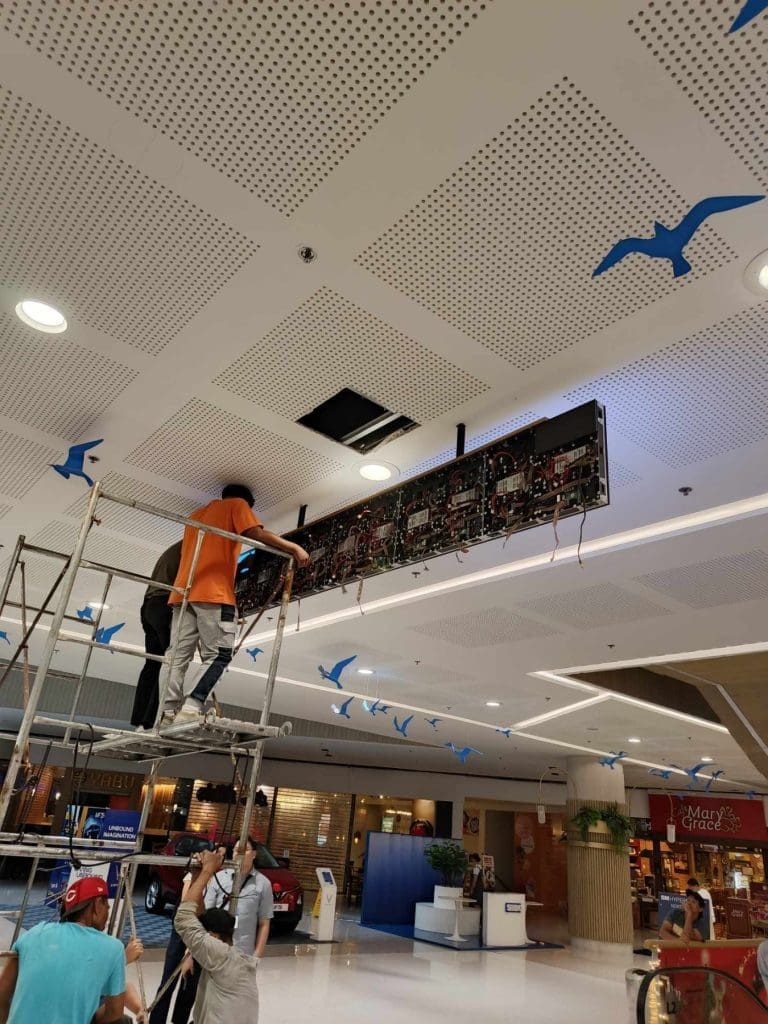 LED signage panel installation on shopping mall ceiling in Quezon City, blue bird decor and shops visible. By M&G Global Ads.