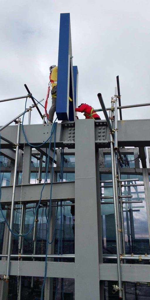 Worker in safety gear installs blue acrylic building signage on scaffolding in Makati, cloudy sky, by M&G Global Ads.
