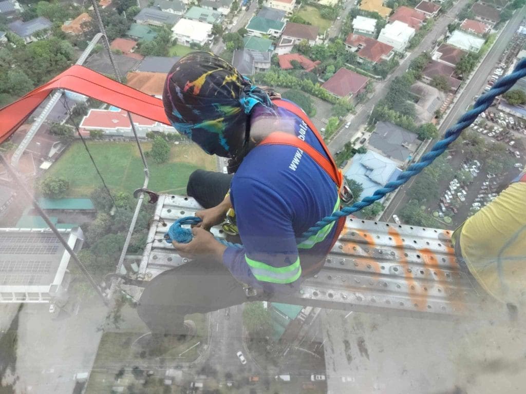 Worker cleans building window on suspended platform beside metallic signage in Ortigas, high above ground level by M&G Global Ads.