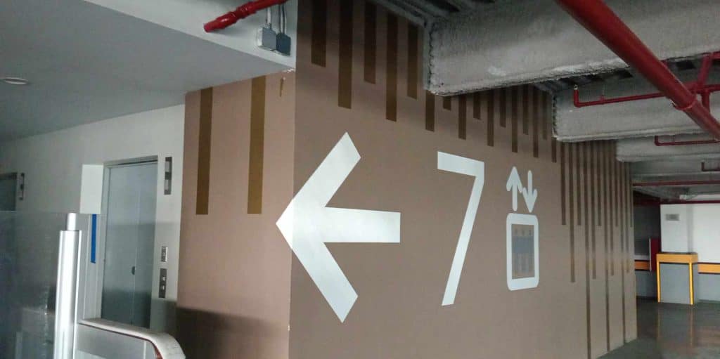 Parking garage directional signage with arrow, 7, and elevator in Makati, metallic sign beside brown wall by M&G Global Ads.