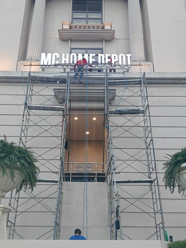 MC Home Depot stainless steel signage installation on building exterior in Pasig, with workers and plants, by M&G Global Ads.