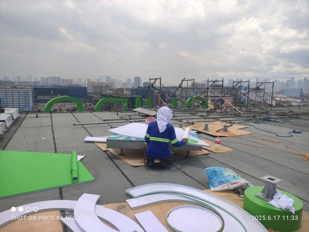 Acrylic 3D signage installation on rooftop in Makati by M&G Global Ads; worker, city buildings, tools, cloudy sky visible.