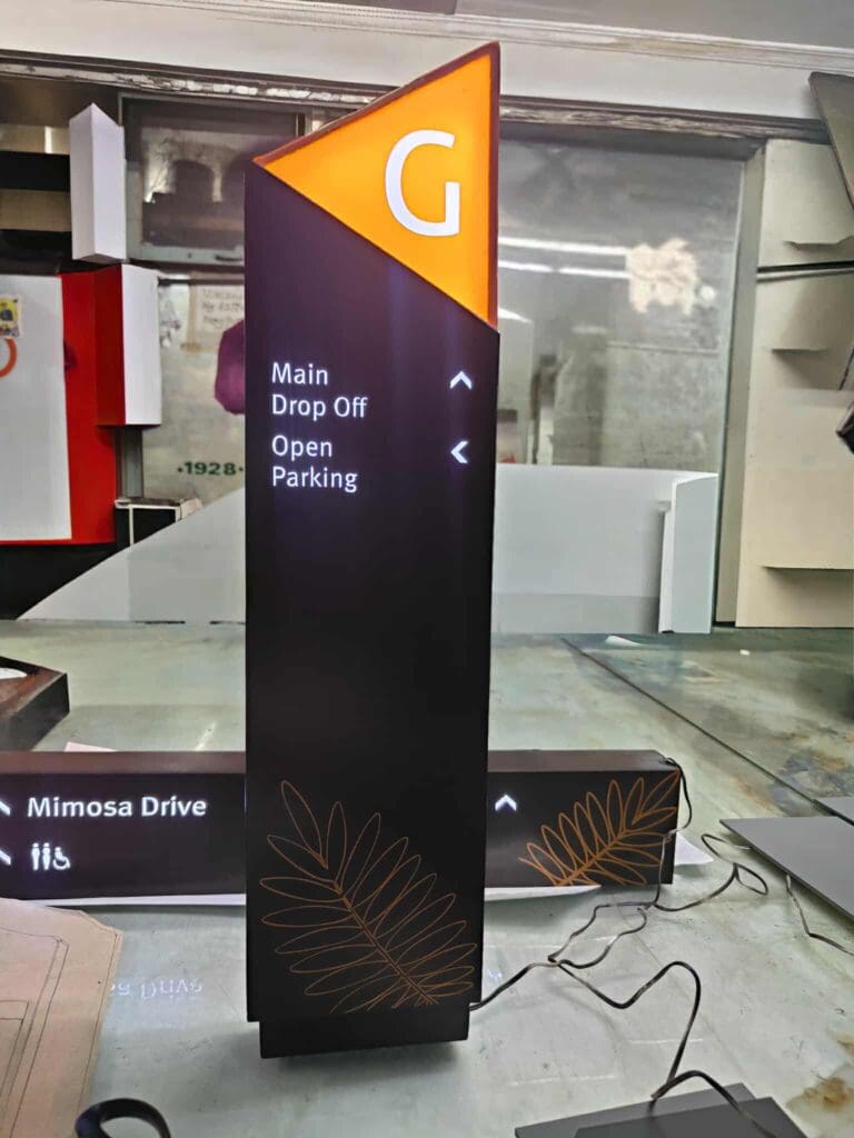 A tall, black directional sign with an orange triangular top labeled G. Below are illuminated white arrows and text reading Main Drop Off and Open Parking. Decorative leaf designs are at the bottom.