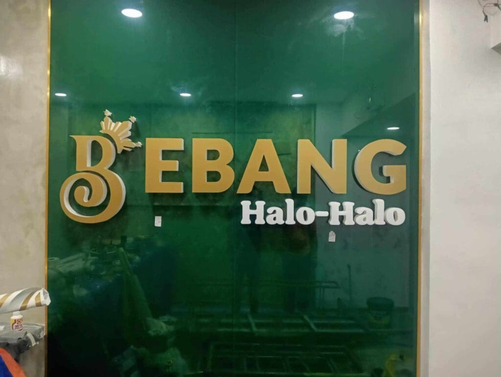 Bebang Halo-Halo gold acrylic signage on green wall in Quezon City, featuring crowned B logo above text by M&G Global Ads.