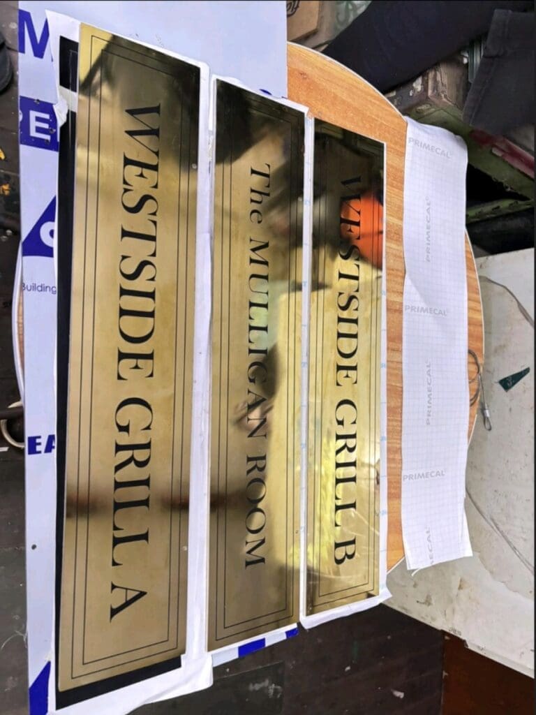 Custom gold metal restaurant door signs for WESTSIDE GRILL in Makati by M&G Global Ads, shown on a wooden surface.
