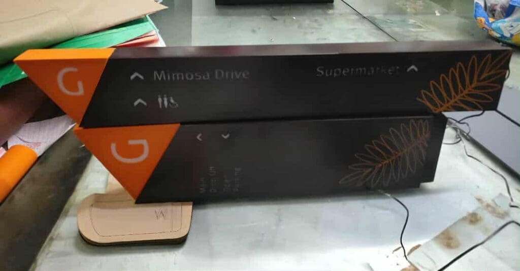 Two black arrow-shaped directional signs with orange accents and leaf designs, mounted on a stand. The top sign points to Mimosa Drive and Supermarket; the bottom sign’s text is partially unreadable.