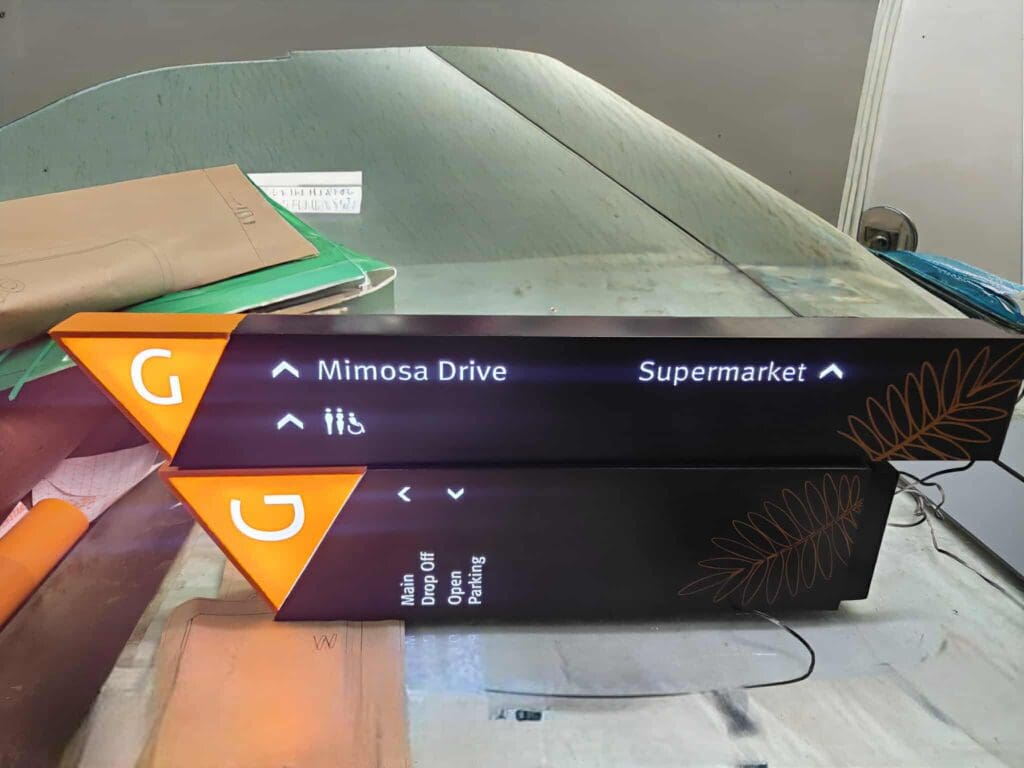 Two illuminated black and orange directional signs with the letter “G.” The top sign points to Mimosa Drive and Supermarket, while the bottom sign has some unreadable text and a list with drop off, open, and parking.