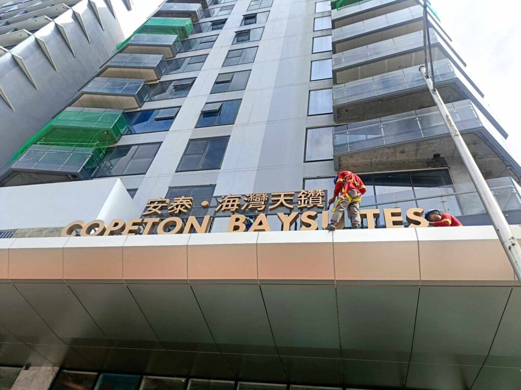Sign maker Philippines installs brass building signage for Copeton Baysuites, Metro Manila, by M&G Global Ads. Two workers at work.