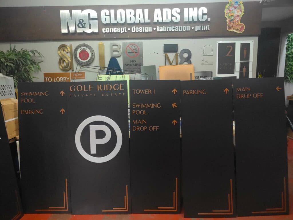 Acrylic and stainless steel wayfinding directional signboards for Golf Ridge Private Estate are showcased in front of the M&G Global Ads Inc. office counter, with multiple signage samples on display behind the counter. Location: Metro Manila, sign maker Philippines by M&G Global Ads.