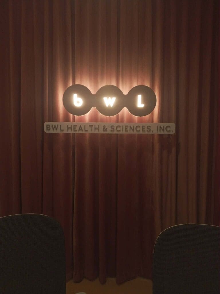 LED acrylic indoor signage for BWL HEALTH & SCIENCES, INC., featuring a glowing "bwl" sign with illuminated dots above an acrylic panel stating the company name, all mounted against a brown curtain background in Metro Manila. Sign maker Philippines by M&G Global Ads.