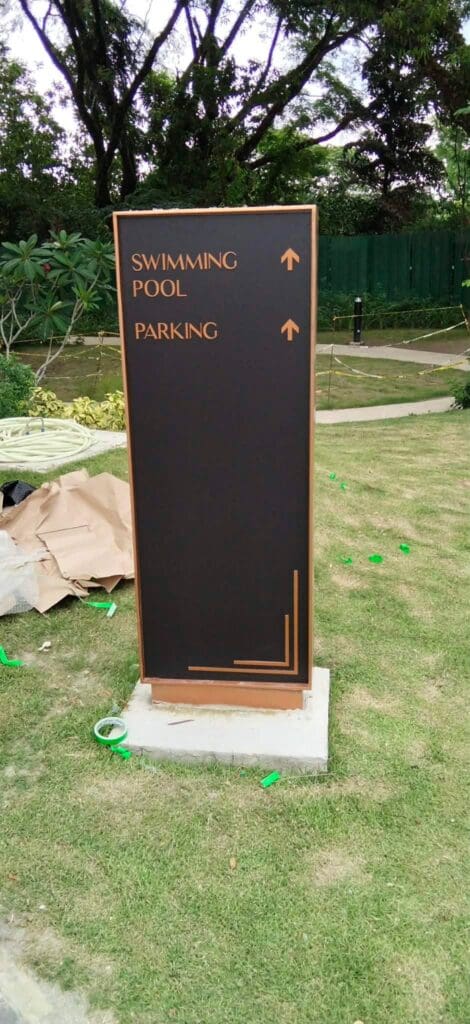 Outdoor wayfinding sign, black and gold, possibly brass or aluminum, with arrows pointing up to indicate directions to the swimming pool and parking area; installed on grass with landscaped greenery and trees in the background in the Philippines. Sign maker Philippines by M&G Global Ads.