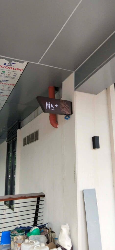Acrylic indoor restroom wayfinding sign with brown background and white icons for men's, women's, and accessible restrooms, plus a plus sign. The sign is mounted under a gray ceiling near a wall with exposed pipes and cleaning supplies visible below. Location: Philippines. Sign maker Philippines by M&G Global Ads.
