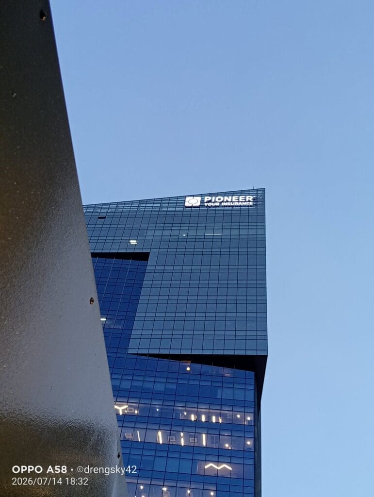 Pioneer Your Insurance acrylic building signage on a modern glass building exterior against a clear blue sky in Metro Manila, viewed from a low angle with part of a metallic structure in the left foreground, sign maker Philippines by M&G Global Ads.