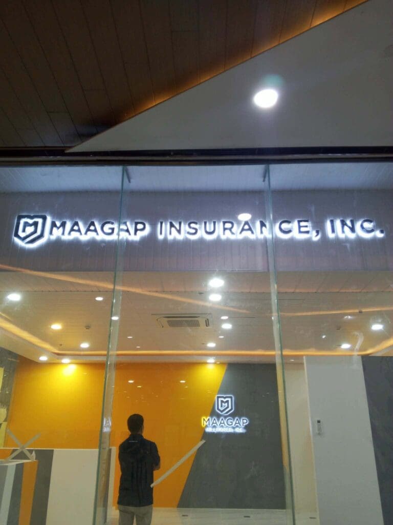 A person stands in front of a glass-walled office with an indoor, brightly lit LED acrylic signage that reads MAAGAP INSURANCE, INC., inside a modern yellow and gray interior. Sign maker Philippines for Maagap Insurance, Metro Manila by M&G Global Ads.