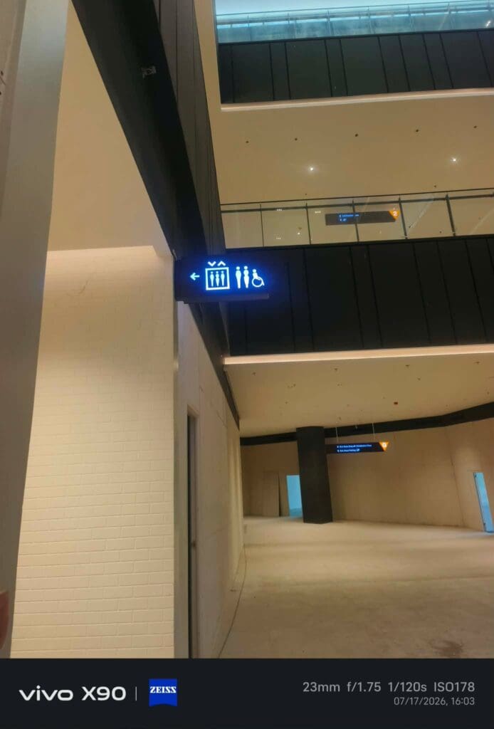 A hallway in a modern building in Metro Manila features indoor wayfinding acrylic signage with icons for a gift shop, restrooms, and accessible facilities. The photo details and timestamp are visible at the bottom. Sign maker Philippines by M&G Global Ads.
