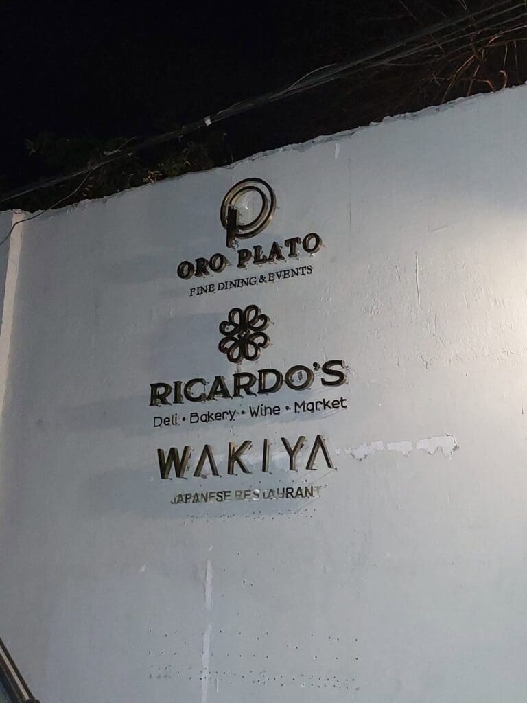 Indoor building signage with gold lettering for Oro Plato Fine Dining & Events, Ricardos Deli-Bakery-Wine-Market, and Wakiya Japanese Restaurant on a white wall, created by sign maker Philippines, Metro Manila location. Signage material features metallic or brass finish. By M&G Global Ads.
