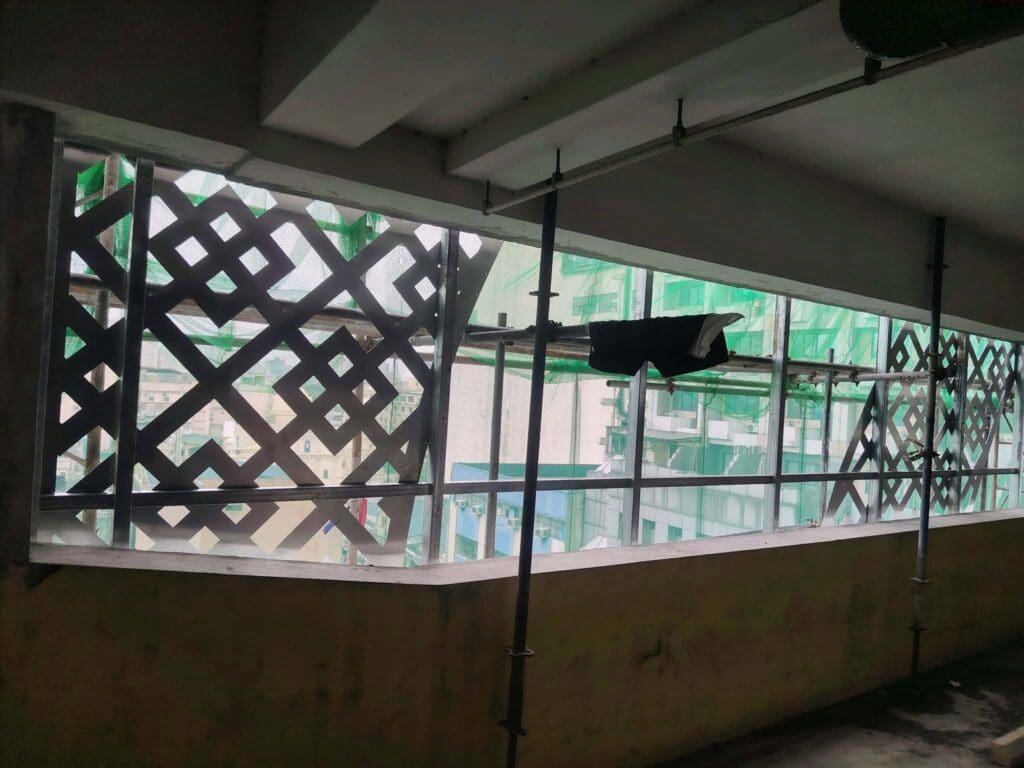 Building signage installation in progress at a partially constructed building in Metro Manila, featuring lattice-patterned window grids, green safety netting, and exterior scaffolding. The unfinished interior includes a piece of black cloth hanging on the railing. Sign maker Philippines by M&G Global Ads.