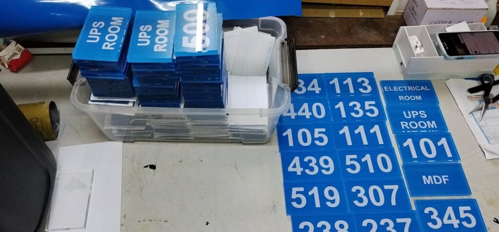 Stacks of blue acrylic indoor room and wayfinding signs, including UPS ROOM, ELECTRICAL ROOM, and various numbers, are arranged neatly on a white tabletop with some stacked in a clear bin and others laid out flat. Sign maker Philippines by M&G Global Ads.