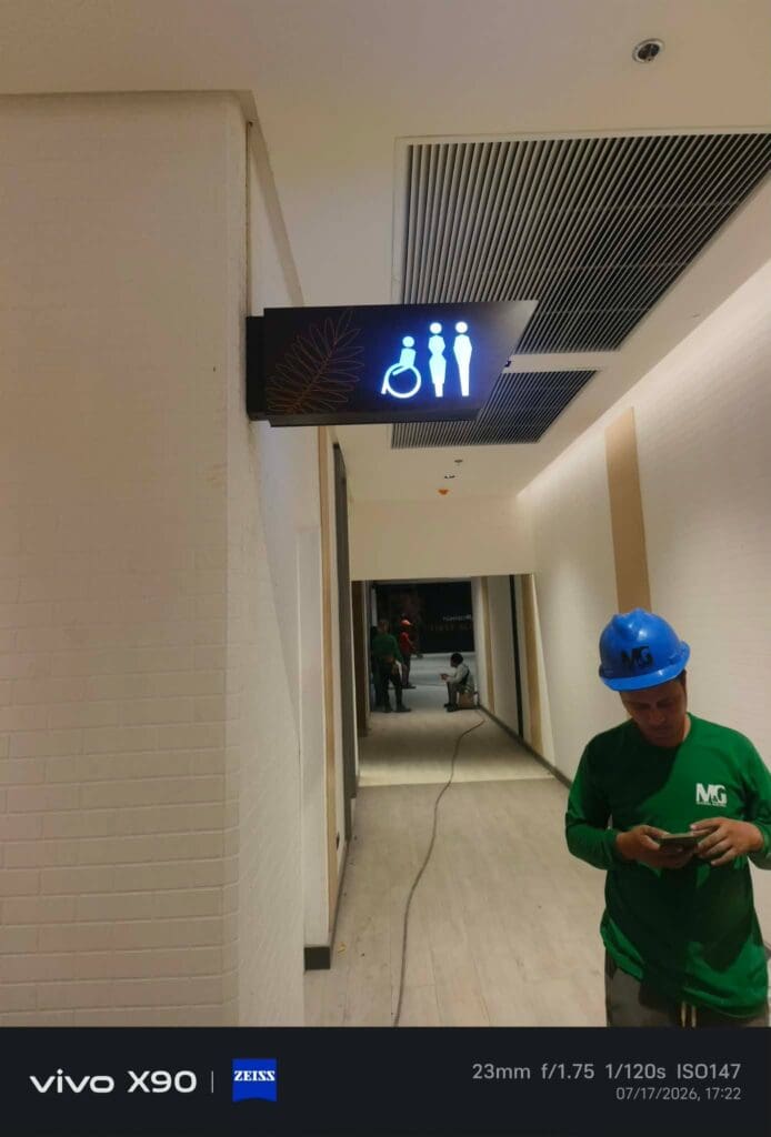 A construction worker in a green uniform and blue helmet stands under an indoor acrylic restroom sign in a hallway with unfinished floors and exposed ceiling vents, Metro Manila. Sign maker Philippines by M&G Global Ads.