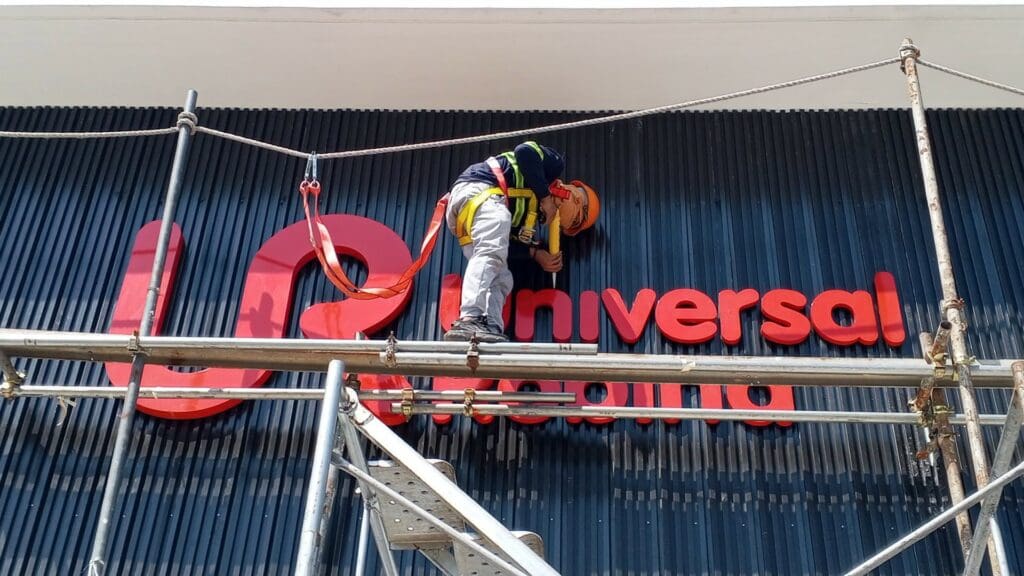 A construction worker wearing safety gear installs or repairs an acrylic red building sign on a blue ribbed wall, standing on scaffolding in front of the word Universal in Metro Manila, sign maker Philippines by M&G Global Ads.
