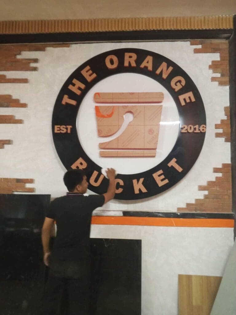 A man cleans a round indoor building acrylic sign for The Orange Bucket, mounted on brick wall, sign maker Philippines by M&G Global Ads.