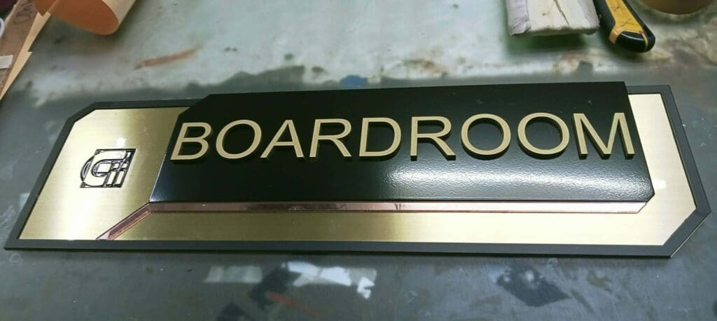 A rectangular black and gold indoor acrylic room sign with raised gold letters spelling BOARDROOM and a logo on the left side, resting on a table. Sign maker Philippines by M&G Global Ads.