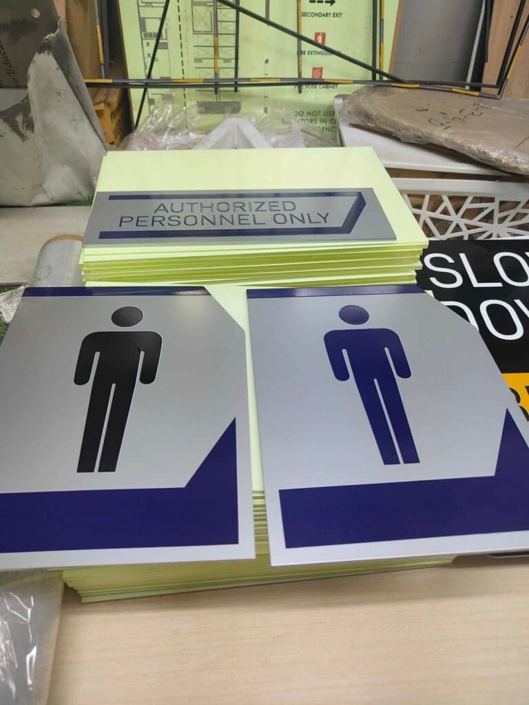 A stack of indoor acrylic safety and restroom signs on a table, including an AUTHORIZED PERSONNEL ONLY sign and two restroom signs with generic human figures in black and blue, sign maker Philippines. Location: Metro Manila. By M&G Global Ads.