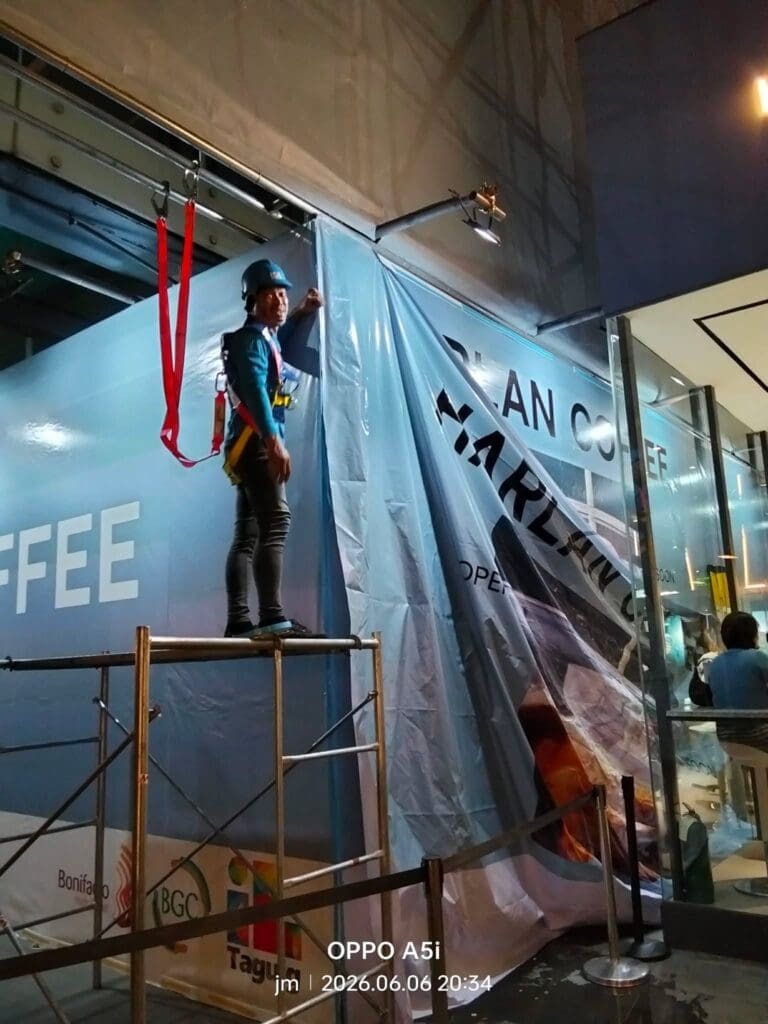 A worker installs a large outdoor printed banner signage on a glass building at night in Metro Manila, sign maker Philippines by M&G Global Ads.