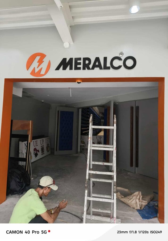 A worker installs an indoor acrylic Meralco building sign above an entrance, unfinished site, Metro Manila, sign maker Philippines by M&G Global Ads.